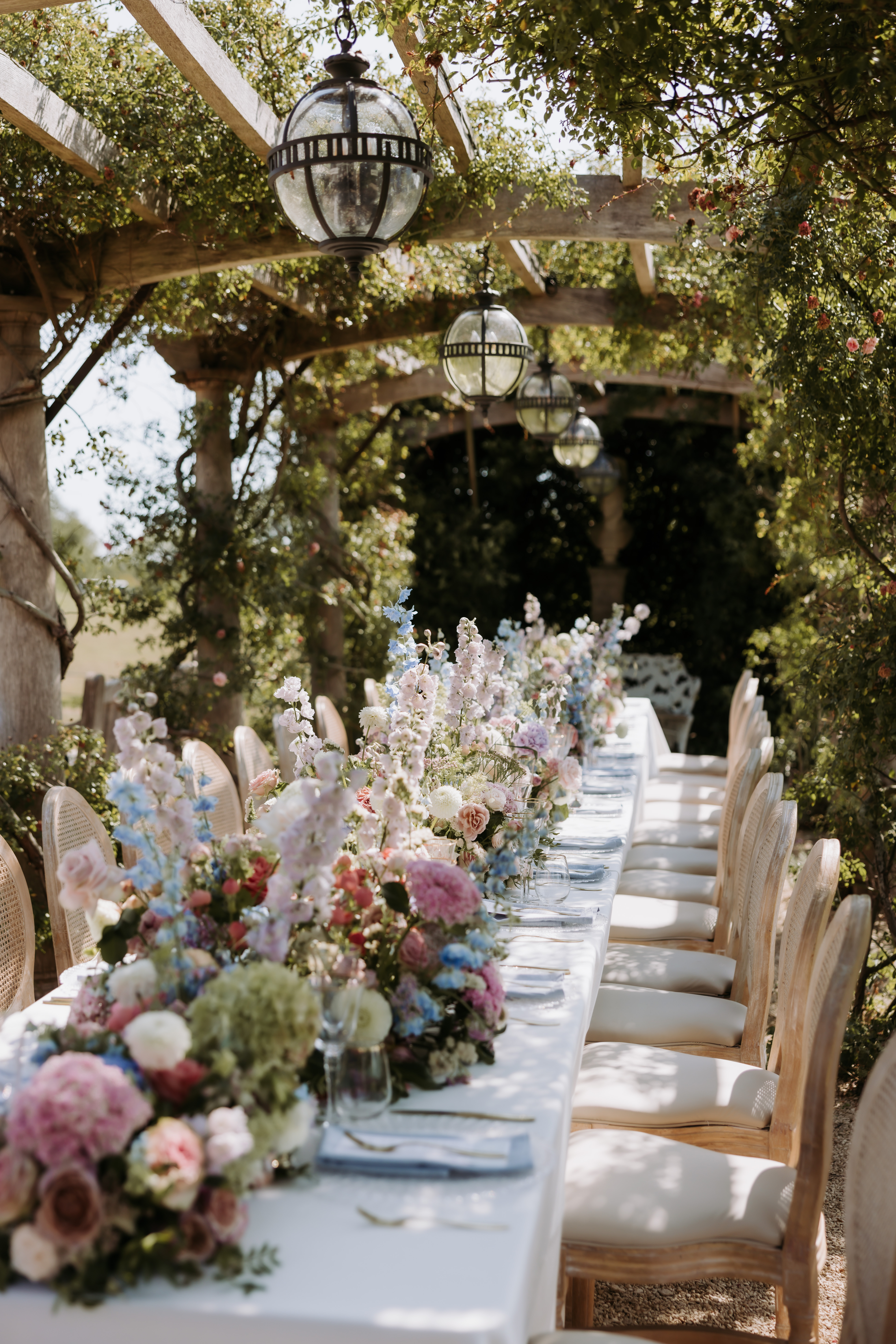 A long wedding table set beneath a pergola with flowering climbers and pendant lanterns